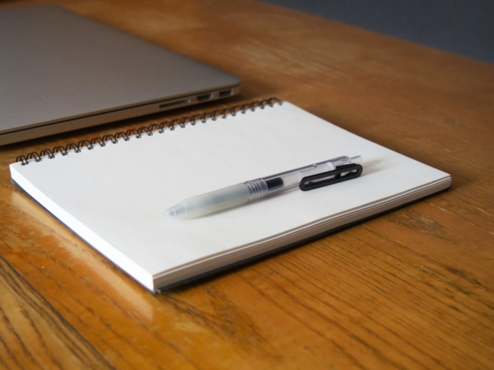 An open spiral notebook and pen on a wooden desk beside a laptop